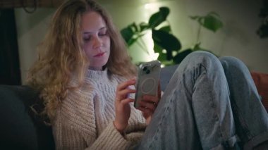 Genç ve güzel bir kadın, bir kış akşamı rahat bir oturma odasında otururken telefonu kullanır.