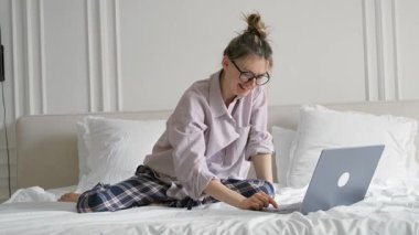 Genç ve güzel bir kadın dizüstü bilgisayarda çalışıyor, sabahları yatağında oturuyor.