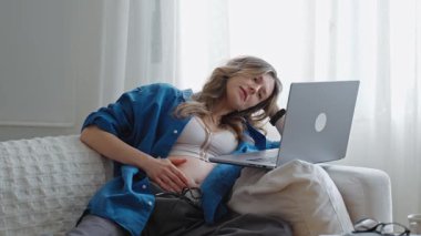 Güzel düşünceli hamile bir kadın oturma odasındaki kanepede otururken dizüstü bilgisayar kullanıyor.