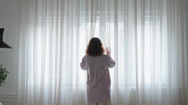Parlak, ferah bir yatak odasında genç bir kadın perdeleri açıyor.