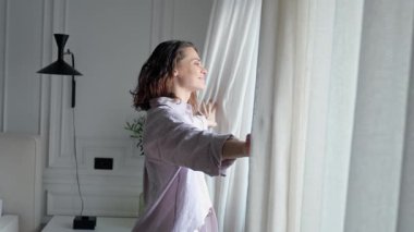 Parlak, ferah bir yatak odasında genç bir kadın perdeleri açıyor.