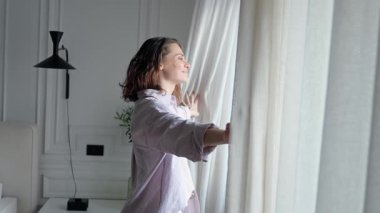 Parlak, ferah bir yatak odasında genç bir kadın perdeleri açıyor.