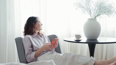 Genç bir kadın, parlak bir oturma odasındaki koltukta otururken sosyal medyada telefonuyla geziniyor.