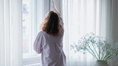 Genç bir kadın perdeleri indirir ve parlak bir oturma odasında pencere açar.