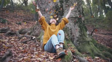 Genç bir kadın sonbahar ormanında büyük bir ağacın yanında otururken düşmüş yaprakları havaya fırlatıyor.