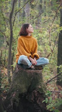 Genç bir kadın sonbahar ormanındaki büyük bir ağacın kütüğünde otururken temiz havanın tadını çıkarıyor.