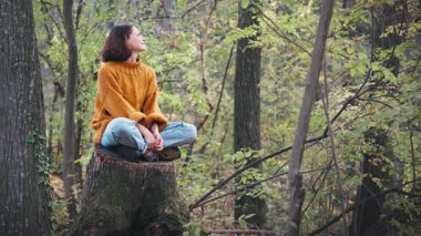 Genç bir kadın sonbahar ormanındaki büyük bir ağacın kütüğünde otururken temiz havanın tadını çıkarıyor.