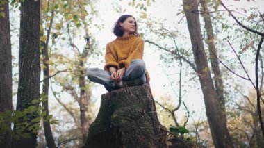 Genç bir kadın sonbahar ormanındaki büyük bir ağacın kütüğünde otururken temiz havanın tadını çıkarıyor.