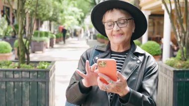 Şık, olgun bir kadın Avrupa 'da bir şehrin caddesinde dururken akıllı telefon kullanır.