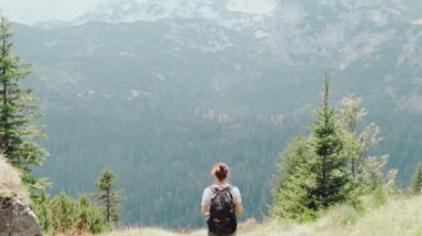 Dağların panoramik manzaralı bir tepenin kenarında duran sırt çantalı genç bir bayan gezgin.