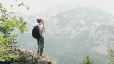 Sırt çantalı genç bir kadın gezgin bir uçurumun kenarında duruyor, dağların panoramik manzarasına hayran.