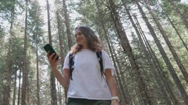 Sırt çantalı genç bir kadın yaz ormanında yürüyüş yaparken akıllı telefonunu kullanıyor.