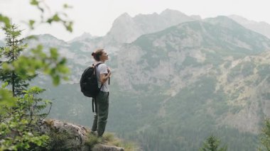 Sırt çantalı genç bir kadın gezgin bir uçurumun kenarında duruyor, dağların panoramik manzarasına hayran.