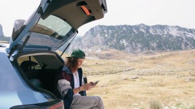 A young woman uses a smartphone while sitting in the trunk of her car with a stunning view of the mountains