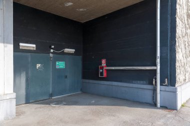 İki gri-mavi çelik garaj kapısı karanlık duvarlı bir binanın köşesinde duruyor. Kapının üstünde iki ışık ve bir güvenlik kamerası var. Kapıların yanındaki duvara kırmızı bir yangın söndürücü yerleştirildi..