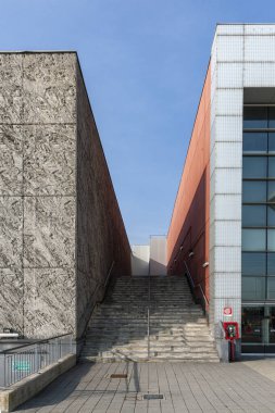 Şehir mimarisi. İki bina arasında taş bir merdiven yükseliyor. Soldaki binanın desenli gri taş bir cephesi var, sağdakinin ise şebeke desenli kırmızı ve beyaz duvarları var..