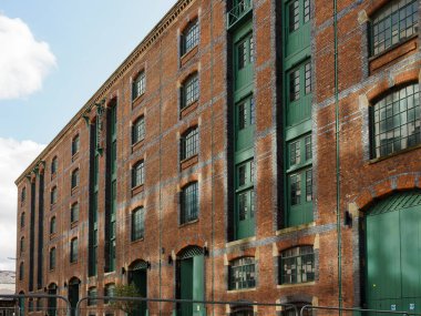 Manchester İngiltere. Parçalı bulutlu gökyüzünün altında yeşil kapıları ve pencereleri olan tarihi tuğla depo..
