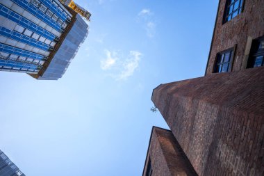 Modern bir cam bina ve tarihi tuğla yapı canlı mavi bir gökyüzüne karşı duruyor. Görüntü, modern ve eski arasındaki mimari karşıtlığı yakalıyor..