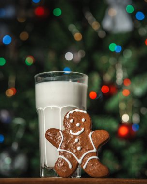Bir bardak sütle zencefilli çörek ve arka planda ışıklarla süslenmiş bir Noel ağacı.
