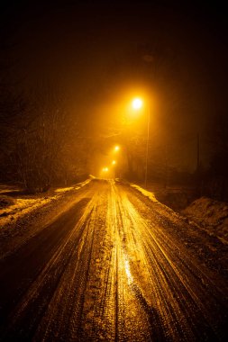 Kırsal bir yolda ıssız bir gece manzarası. Yol karla kaplı ve ıslak görünüyor, sokak lambalarından gelen sıcak turuncu ışığı yansıtıyor. Etrafı sislerle çevrili..