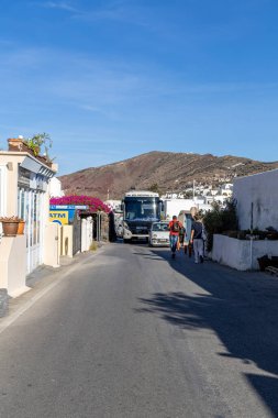 Santorini, Yunanistan - 15 Ekim 2024: Oia 'da bir tur otobüsü geçerken ve yayalar arka planda geleneksel bembeyaz binaların ve canlı bougainvillea' nın önünden geçerken işlek ana cadde