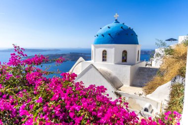 Geleneksel Santorini mavi kubbeli kilise pembe begonvillerle çevrili ve Ege Denizi manzarası nefes kesici.