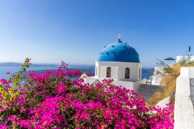 Santorini 'de güneşli bir günde okyanusa bakan mavi kubbeli ve pembe çiçekli geleneksel Yunan beyaz kilisesi
