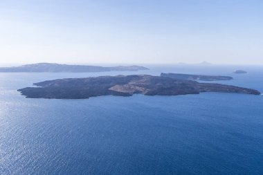 Santorini Caldera 'daki Nea Kameni volkanik adasının havadan görüntüsü. Etrafı koyu mavi Ege Denizi ile çevrili. Parlak yaz gökyüzü ve uzak ada siluetleri altında..