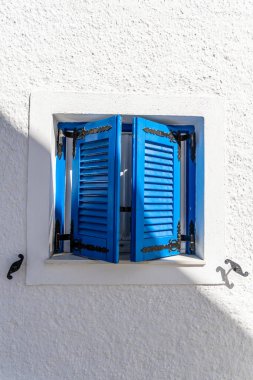 Güneşli bir Cycladic penceresinin ayrıntıları. Canlı mavi panjurlar ve süslü metal menteşeler. Huzurlu bir Santorini köyünde beyazlatılmış bir duvarda..