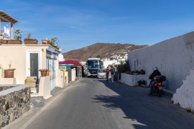 Santorini, Yunanistan - 15 Ekim 2024: Oia 'nın canlı ana caddesi dar yollardan geçen, beyaz binalar, dükkanlar ve açık mavi gökyüzü altında çiçek açan bir otobüsle
