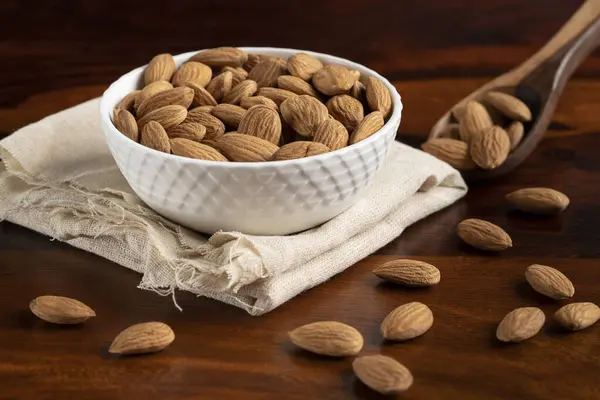 A white bowl full of organic almonds kept on white cotton cloth and almonds spread around wooden spoon