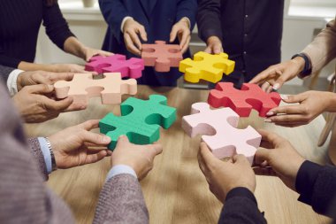 İş ekibi renkli yapboz parçalarını eşleştirmeye çalışıyor. Tahta masanın etrafında duran bir grup genç insan ellerinde kırmızı, sarı, yeşil, pembe yapboz parçaları tutuyorlar. Takım çalışması konsepti