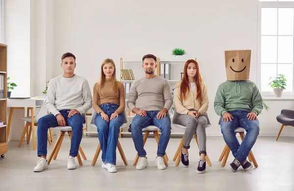 Diverse group of people sits together in row, united as a team. One person wears a paper bag over head, adorned with a hand drawn smile and happy facial expression, creating a whimsical expression.