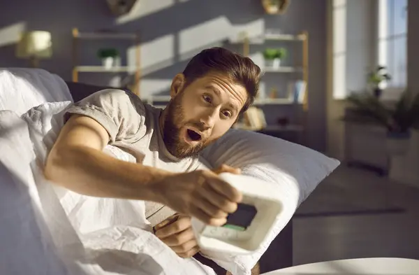 Emotional shocked overslept young redhead man holding alarm clock in his hand, waking up too late for work lying in bed at home. Oversleeping and bad morning concept. Selective focus