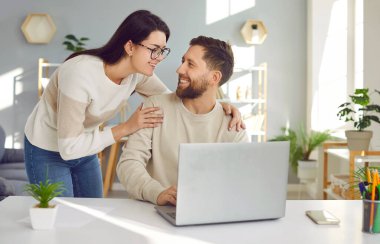 Dizüstü bilgisayarlarını birlikte kullanan mutlu, genç, huzurlu aile çifti. Kocasının çalışma masasında bilgisayarla otururken gülümseyerek kocasını omuzlarından tutan sevgi dolu bir kadın.