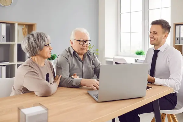 Smiling happy elderly couple talking with man advisor about health insurance or investments sitting at the desk in office with laptop. Senior man and woman talking with male financial agent.