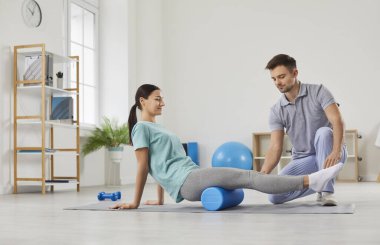 Genç, gülümseyen kadın hasta rehabilitasyon kliniğindeki erkek fizyoterapist doktorun desteğiyle spor masaj silindiri kullanıyor. Fizyoterapi ve fizik tedavi konsepti.