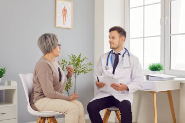 Tıp bürosundaki yaşlı, gri saçlı bir kadının portresi elinde randevu ile rapor tutan ve klinikteki tıbbi muayene sırasında danışmanlık yapan bir erkek doktorla konuşuyor..