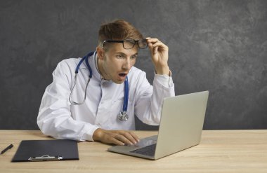 Şok olmuş ve kafası karışmış bir şekilde utanmış erkek doktor dizüstü bilgisayar ekranına bakıyor. Yüzünde korku ifadesi olan bir adam gözlüğünün altından tıbbi bir hataya bakıyor. Gri arkaplan.