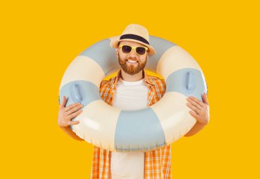 Yazlık giysiler içinde gülümseyen beyaz bir adamın portresi, elinde şişme bir yüzük, turuncu bir arka planda kameraya gülümsüyor. Yaz tatilinin ve sahil seyahatinin neşesini temsil ediyor..