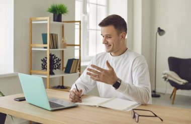 Mutlu bir adam çalışma masasında oturuyor, modern bir dizüstü bilgisayar kullanıyor, video aracılığıyla iş görüşmesi yapıyor, iş arkadaşıyla konuşuyor, ajandasını tartışıyor ve ajandasını kontrol ediyor.