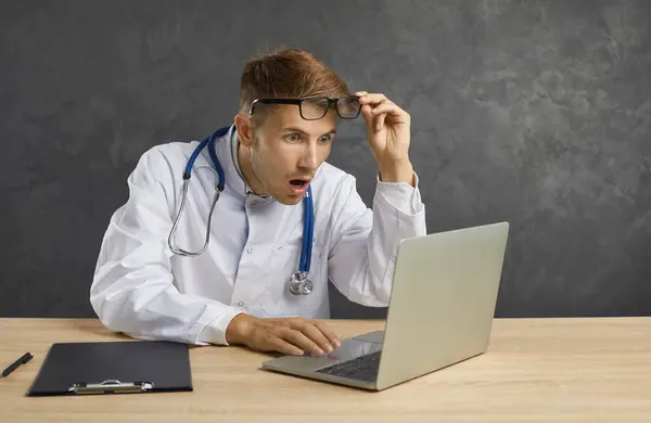 Embarrassed male doctor with a shocked and confused face looks at the laptop screen. Man with a frightened expression looks out from under his glasses looking at a medical error. Gray background.