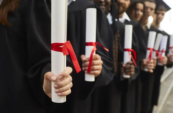 Hands of a graduate students standing in a row in black robes with diplomas in their hands outdoor at graduation ceremony close-up, selective focus. Graduating from university or college concept.
