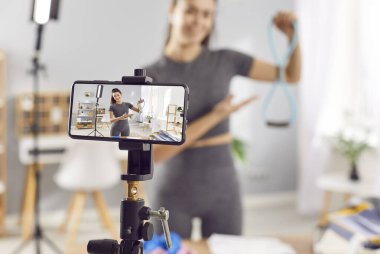 Spor Slim Genç kadın eğitmeni veya spor giyim blogunda online eğitim hakkında video kaydı yapan spor yazarı tripod üzerinde telefon kamerası kullanarak lastik bantlı spor egzersizleri hakkında konuşuyor.