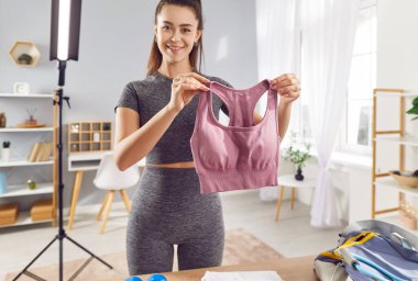 Sportif gülümseyen genç bayan fitness eğitmeni video kaydediyor ve spor kıyafetlerini spor çantalarını hazırlarken gösteriyor. Kız spor blogcusu akıllı telefondaki fitness blogu için video hazırlıyor.