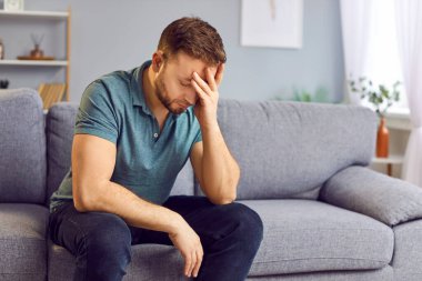 Üzgün, depresif bir adamın portresi. Oturma odasındaki kanepede tek başına oturan baş ağrısından muzdarip. Kötü düşünceler içindeki genç adam evde yalnız hissediyor ya da ilişkilerinde sorunlar yaşıyor.