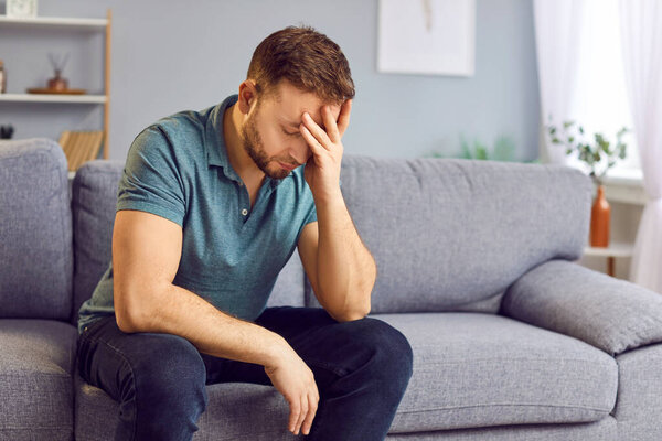 Portrait of upset sad depressed man sitting on sofa in living room at home alone suffering from headache. Young guy in bad thoughts feeling lonely indoors or having problems in relationships