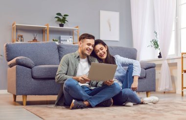 Genç, neşeli bir çift evdeki oturma odasında dizüstü bilgisayarlarını internet ve sosyal medya olarak kullanıyor. Mutlu adam ve kadın kanepede dinleniyor. Haftasonu bilgisayarda video izliyorlar..