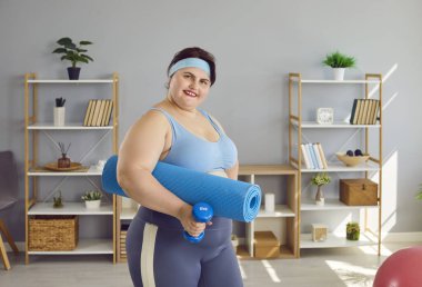 Genç, komik şişman kadın spor egzersizleri yapacak, yoga minderi ve dambılı evde tutacak. Ayrıca kendine güvenen şişman bir kız kameraya bakıyor ve gülümsüyor. Spor ve spor konsepti.