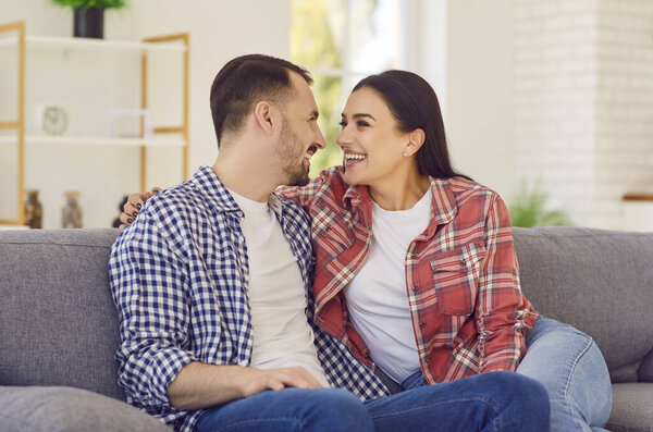 Портрет молодой счастливой влюбленной супружеской пары, сидящей дома на диване. Улыбающиеся семьянин и женщина на диване смотрят друг на друга. Концепция гармонических отношений.